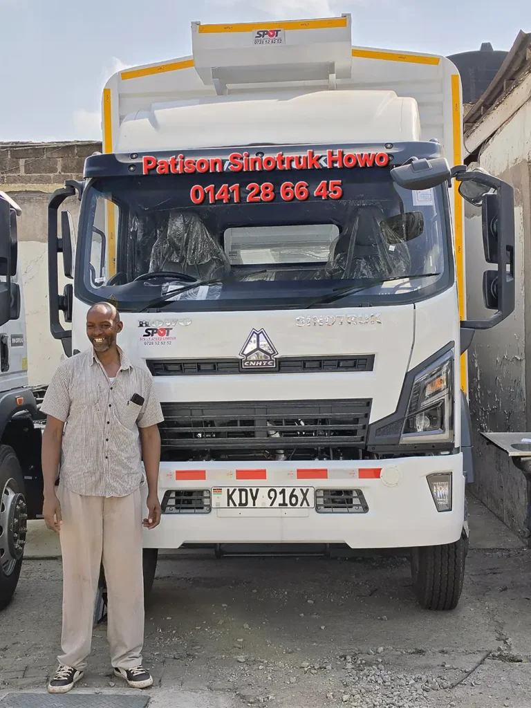 Handover of a new white Sinotruk Howo M3 Rigid 5200MM, registration KDY 916X, to Kabu Plast Ltd at the Patison Sinotruk Howo yard in Nairobi - a smiling man in a checked short-sleeved shirt stands beside the front of the truck, which carries a tall white high-sided cargo body with yellow trim, with the seats still in their factory plastic and another Howo parked alongside